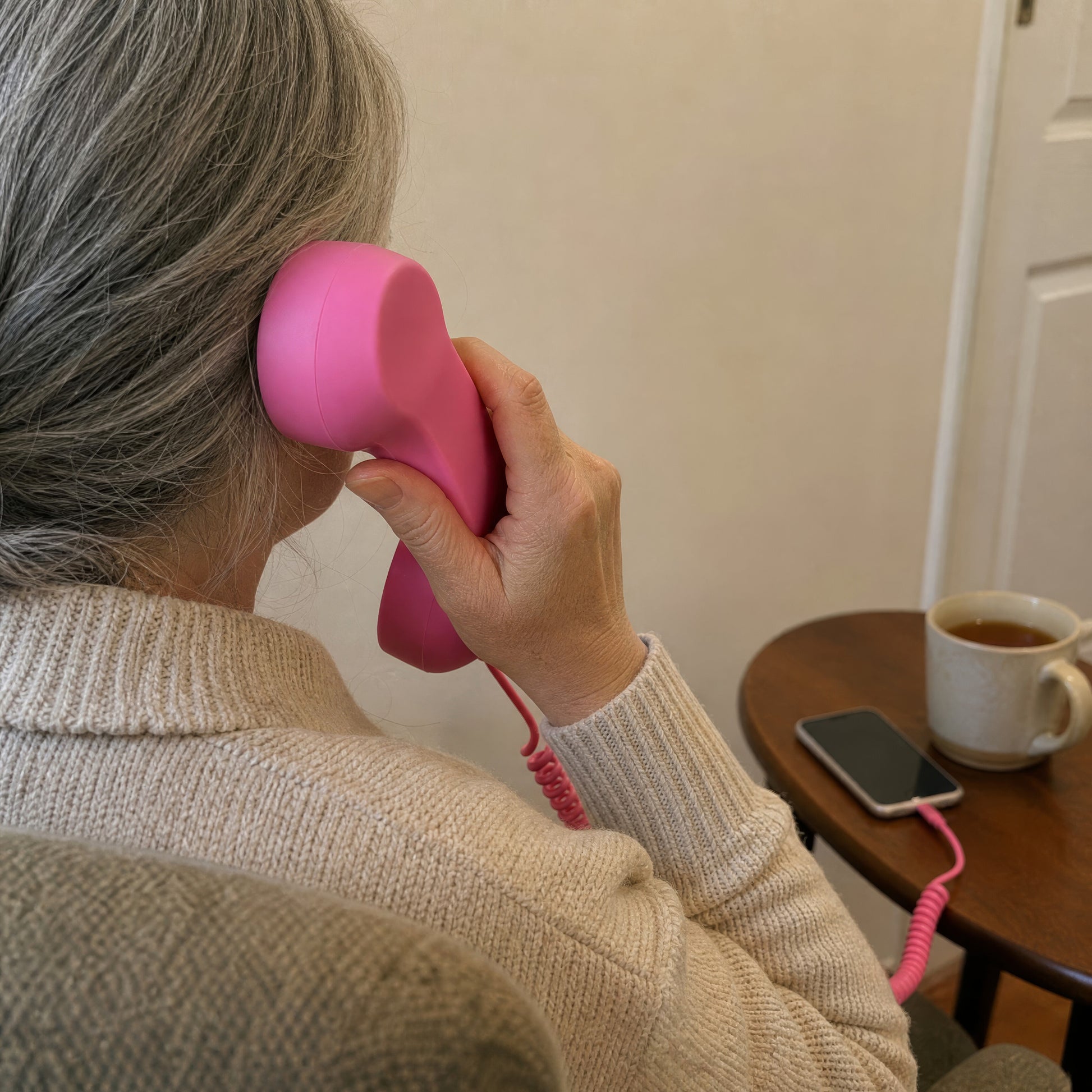 A woman in an armchair holds the pink DialBop handset to her ear, cord running to her phone on the side table