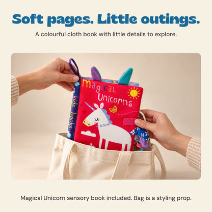Hands hold the red Magical Unicorn cloth sensory book above a plain tote bag, showing its exact cover, loop and fabric tabs. The tote is not included.